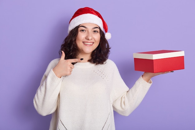 Brunetka pokazuje prezent świąteczny w czerwonym pudełku, wskazując palcem wskazującym, dama w czapce Mikołaja i białym swetrze, dziewczyna na liliowej ścianie, faliste włosy kobieta o szczęśliwym wyglądzie.