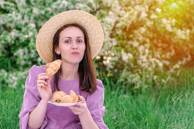 Brunetka w fioletowej sukience i słomkowym kapeluszu je na pikniku świeże rogaliki