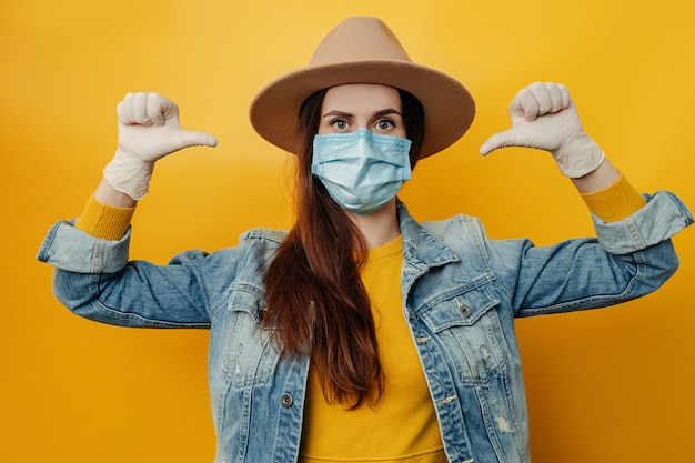 Zdjęcie brunetka w kapeluszu nosi ochronną maskę medyczną, aby nie zarażać innych ludzi, wskazując na siebie kciukami, patrzy w kamerę, na białym tle na żółtym tle. koncepcja koronawirusa, ochrona przed wirusami