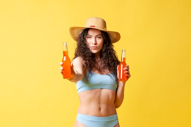 Brunetka w niebieskim kostiumie kąpielowym trzyma butelki z pomarańczowym napojem i oferuje napoje na żółtym tle
