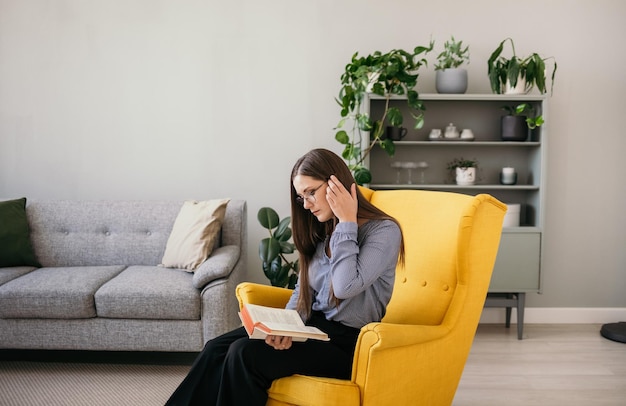 Brunetka w okularach i niebieskiej bluzce siedzi w żółtym fotelu w pokoju i czyta książkę
