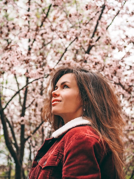 Brunetka z długimi włosami w czerwonej kurtce otoczona różowymi kwiatami Sakury