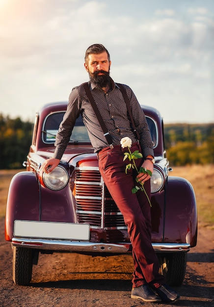 Brunetka z gęstą czarną brodą w koszuli, spodniach na szelkach trzyma w dłoni białą różę w pobliżu brązowego samochodu retro na wiejskiej drodze.