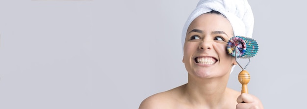 Brunetka z ręcznikiem na głowie używająca wałka do masażu twarzy na szarym tle