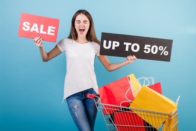 Zdjęcie brunetki kobieta z pushcart zakupy pełno kolorowe czerwone i żółte papierowe torby i 50% sprzedaży znak odizolowywający nad błękitem