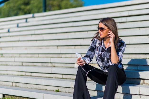 Brunetki kobiety obsiadanie na schodkach podczas gdy słuchający muzykę i używać smartphone