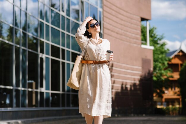 Brunetki Młoda Kobieta Z Okularami Przeciwsłonecznymi I Torbą Trzymającą Kawę Chodzenie Po Mieście. Styl życia Portret Kobiety