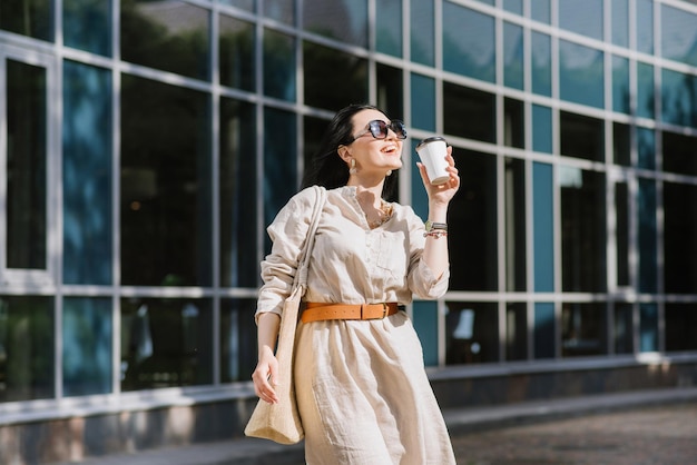 Brunetki młoda kobieta z okularami przeciwsłonecznymi i torbą trzymającą kawę chodzenie po mieście. Styl życia portret kobiety