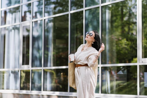 Brunetki młoda kobieta z okularami przeciwsłonecznymi i torbą trzymającą kawę chodzenie po mieście. Styl życia portret kobiety