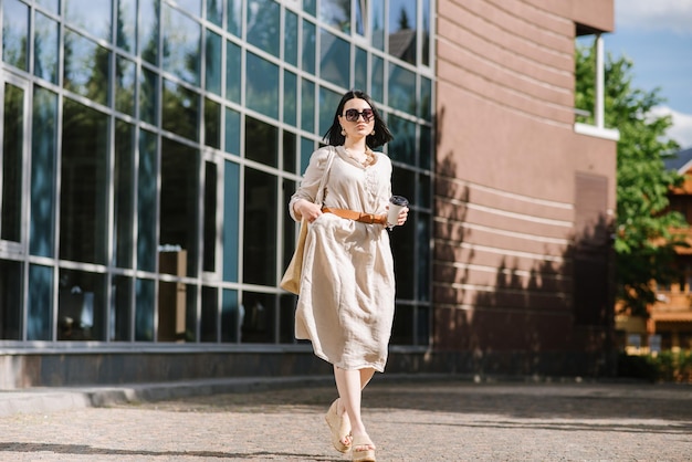 Brunetki młoda kobieta z okularami przeciwsłonecznymi i torbą trzymającą kawę chodzenie po mieście. Styl życia portret kobiety