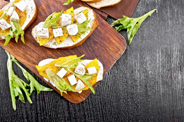 Bruschetta z pieczoną dynią, solonym serem feta, ricottą, rukolą i przyprawami, ręcznik na tle drewnianej deski z góry