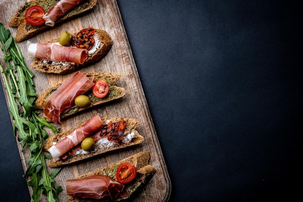 Bruschetty z jamonem, oliwkami, pesto, grillowanymi pomidorkami koktajlowymi podawane na drewnianej desce z rukolą na tle z miejscem na kopię. Śródziemnomorski chleb tostowy z mięsem, serem i warzywami