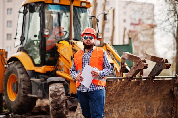 Brutalny broda pracownik człowiek garnitur robotnik budowlany w bezpieczeństwo kask pomarańczowy, okulary przeciw traktor z planem papieru pod ręką.