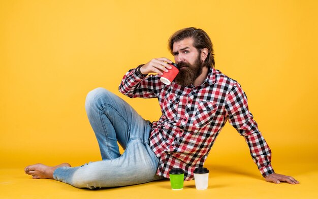 Brutalny brodaty mężczyzna nosi kraciastą koszulę z bujną brodą i wąsami pije poranną kawę z papierowego kubka barista