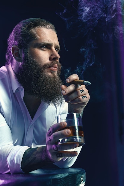 Zdjęcie brutalny brodaty mężczyzna pali cygaro pije whisky dym z dymem ubrany w białą koszulę strzał studyjny