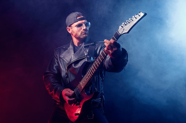 Brutalny brodaty muzyk heavymetalowy w skórzanej kurtce i okularach przeciwsłonecznych gra na gitarze elektrycznej na czarno