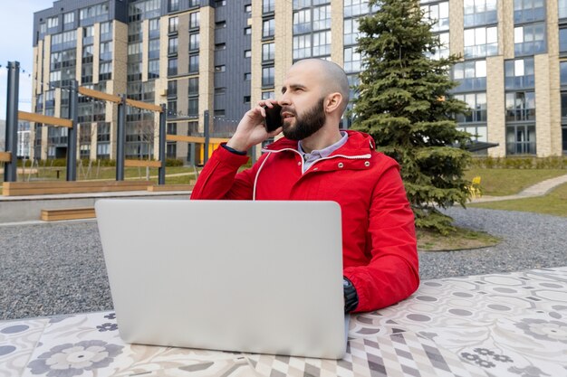 Brutalny Facet Z Brodą W Czerwonej Kurtce Pracuje Przy Komputerze I Rozmawia Przez Telefon Na Ulicy. Koncepcja Zatrudnienia Online