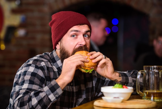 Brutalny hipster brodaty mężczyzna siedzi przy liczniku baru Wysokokokaloryczne jedzenie Podstępny posiłek Pyszna koncepcja hamburgera Cieszyć się smakiem świeżego hamburgera Hipster głodny mężczyzna je hamburger Mężczyzna z brodą je menu hamburgera