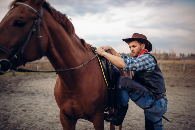 Brutalny kowboj w dżinsach i skórzanej kurtce wspina się na koniu na ranczo w Teksasie, western. Vintage mężczyzna z koniem, dziki zachód