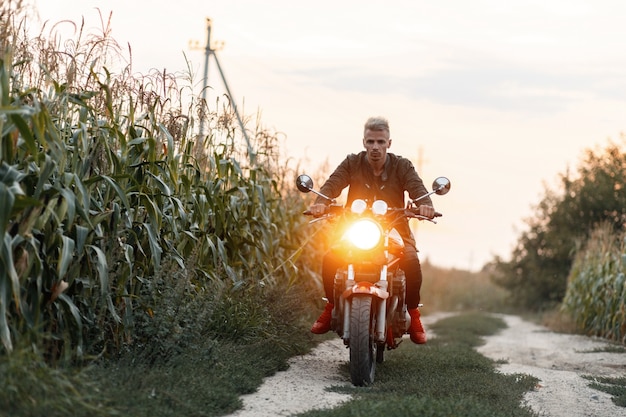 Brutalny mężczyzna w wojskowej kurtce jeździ motocyklem ze światłem po polu kukurydzy