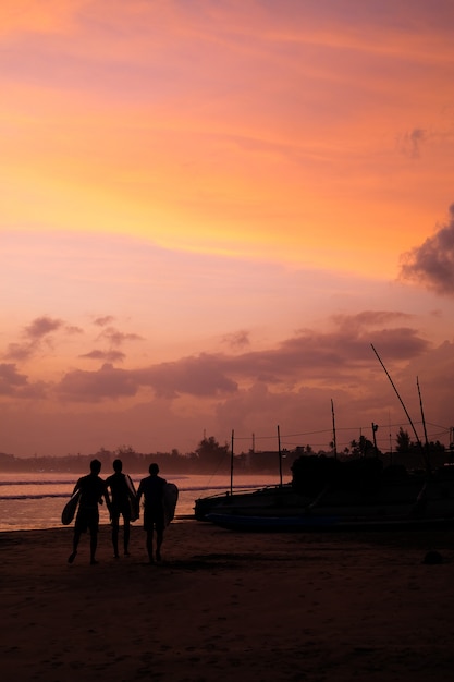 Zdjęcie brzeg oceanu o zachodzie słońca sylwetki łodzi i surferów ludzi iść na plażę