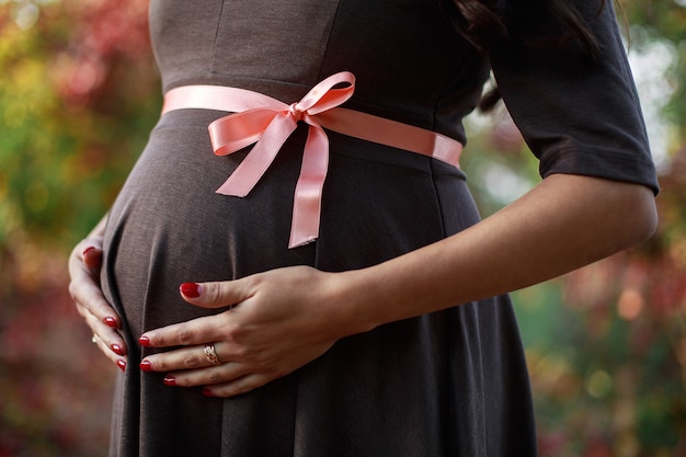 Brzuch kobiety w ciąży związany różową wstążką. piękna kobieta w ciąży czule obejmując jej brzuch w ciąży z bliska. Portret ciężarnego delikatnego macania ciężarny brzuch na naturze.