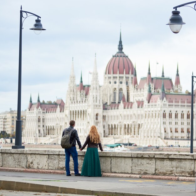 Budynek Parlamentu w Budapeszcie, Węgry. Widok z tyłu pary turystów trzymających się za ręce i cieszących się widokiem wspaniałej architektury Budapesztu między dwiema starożytnymi latarniami