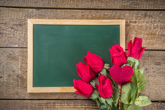 Bukiet czerwonych kwiatów róż z czerwonym sercem na drewnianym stole z tablicą. Walentynki wakacje