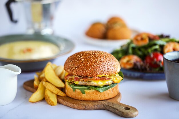 Burger z frytkami wśród jedzenia. Burger z Wołowiną, Serem, Bekonem. Lunch na stole Zbliżenie hamburgera z sałatką, ser na desce Klasyczny burger Dzień posiłków Jedzenie na stole Pyszne rupiecie