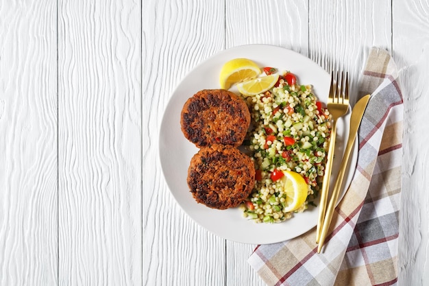 Burgery jagnięce Harissa podane z sałatką z kuskusu perłowego na talerzu z plastrami cytryny na białym drewnianym stole ze złotymi sztućcami, widok poziomy z góry, leżący płasko