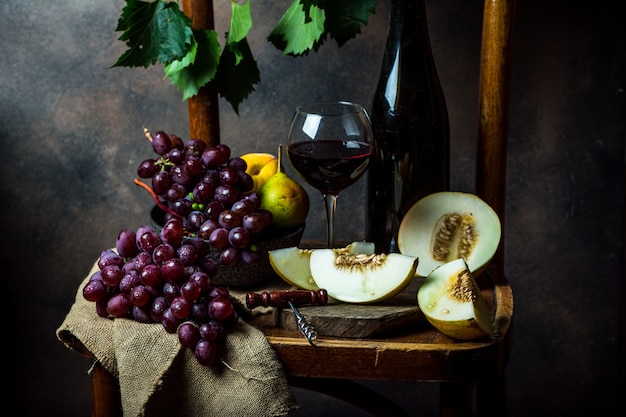 Butelka i kieliszek czerwonego wina, winogron i korka na krześle. Melon, kawałek melona. Różowe winogrono, gruszka. Martwa natura z jedzeniem. Fotografia ciemnych potraw. Koncepcja jesień. Alkohol, uprawa winorośli.