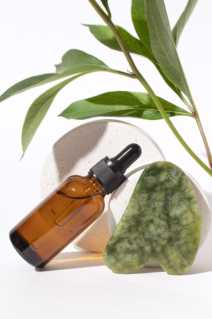 Butelka kosmetyczna z zakraplaczem na podium Jade Jade Gua sha do masażu twarzy Pojemnik na serum i gałązki roślin na białym tle Koncepcja produktów kosmetycznych kosmetyków organicznych