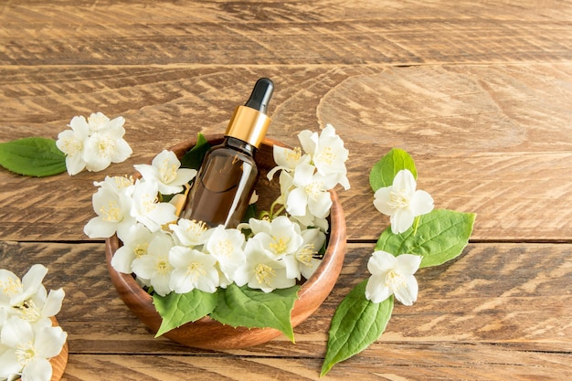 Butelka kosmetyczna z zakraplaczem z ciemnego szkła z olejkiem organicznym leży w drewnianej miseczce wśród kwiatów pachnącego jaśminu kosmetyków naturalnych