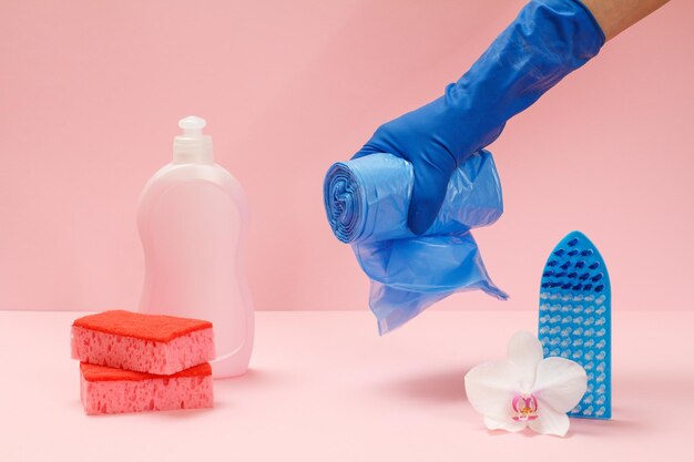 Butelka płynów do mycia naczyń, gąbki, szczotka i ręka w gumowej rękawiczce trzymająca niebieskie worki na śmieci