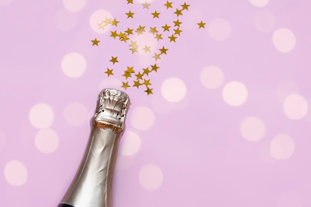 Butelka szampana z konfetti złotej gwiazdy na pastelowym różowym tle i bokeh świateł z copyspace