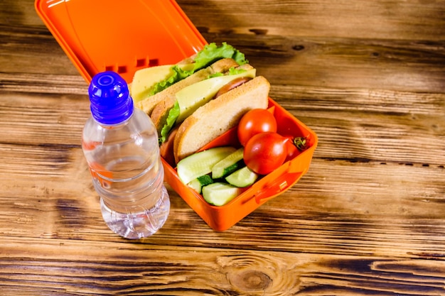Butelka wody i pudełko na lunch z kanapkami, ogórkami i pomidorami na rustykalnym drewnianym stole