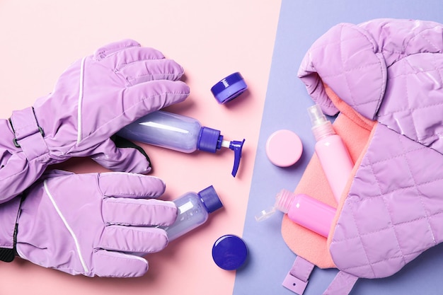Butelki kosmetyczne i słoiki z nakrętką i rękawice narciarskie na różowym i fioletowym tle widok z góry