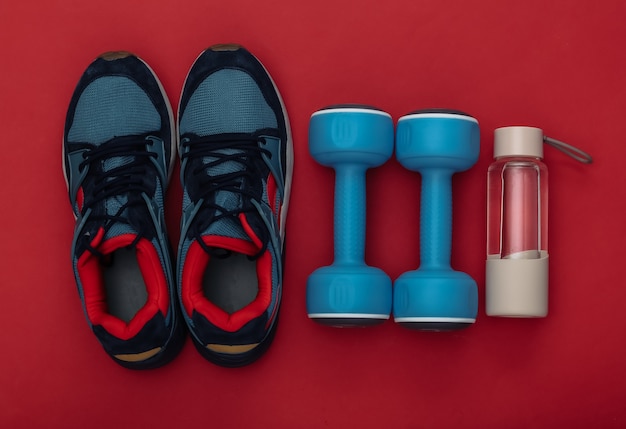 Buty sportowe (trampki) i niebieskie hantle, bidon na czerwonym tle. Zdrowy styl życia, trening fitness. Widok z góry