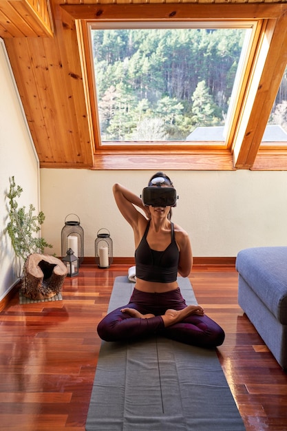 Całe ciało bosej kobiety w okularach wirtualnej rzeczywistości i siedzącej w pozie Lotosu ze skrzyżowanymi nogami podczas ćwiczenia medytacji jogi w domu