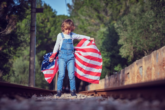 Całe ciało nastoletniego chłopca z flagą Stanów Zjednoczonych Ameryki stojącego na opuszczonej linii kolejowej i patrzącego w dół