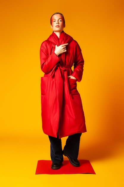 Całe ciało szczupła kobieta z kreatywnym makijażem i stylowym workowatym płaszczem, stojąc na kartce czerwonego papieru na żółtym tle