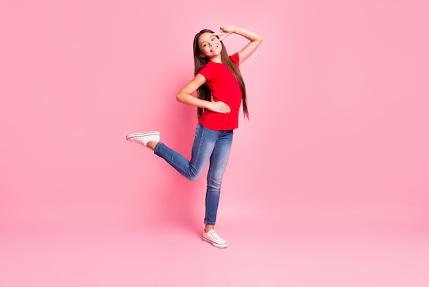 Całe zdjęcie wielkości ciała ślicznej słodkiej małej damy długiej fryzury pokaż znak v taniec podnieś nogę ręka biodra ząb uśmiech nosić dorywczo czerwony t-shirt dżinsy trampki na białym tle różowy kolor tła