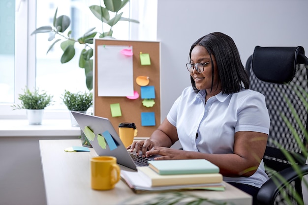 Całkiem czarna dama pisania na klawiaturze laptopa działa online w przytulnym, nowoczesnym biurze. Amerykanka z czarnymi włosami i skórą lubi pracować sama, zarządzać. założyć działalność. w miejscu pracy