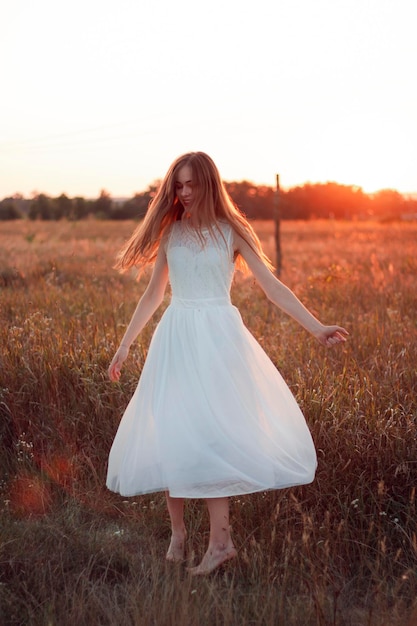 Całkiem ładna dziewczyna krąży w białej sukni na polu. Promienie słoneczne. Pomarańczowy zachód słońca. Pojęcie. Kobieta