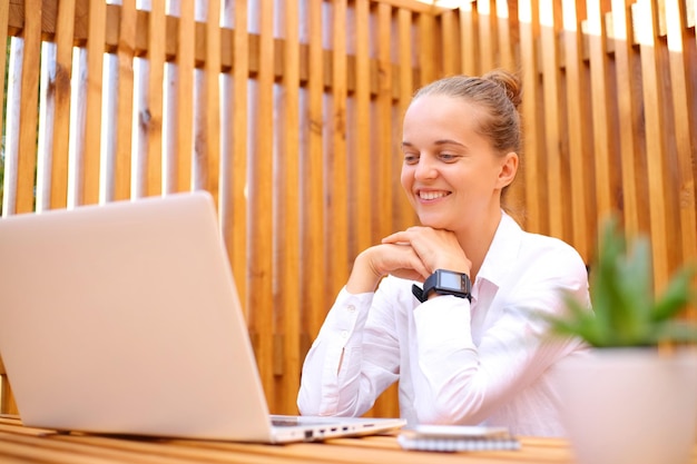 Całkiem ładna kobieta ubrana w białą koszulę, siedząca w kawiarni na świeżym powietrzu, pracująca na laptopie, mająca konferencję online, która spotyka się z pozytywnymi emocjami