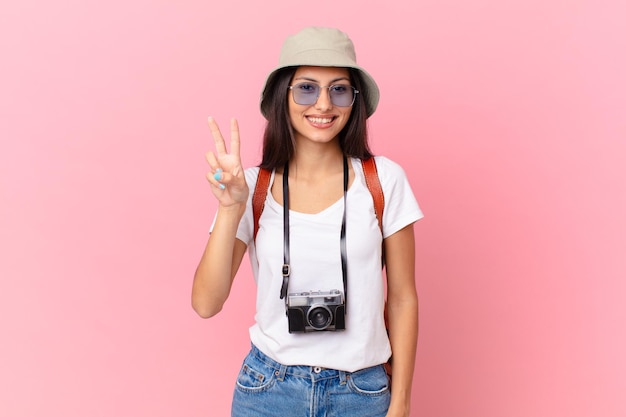 Całkiem latynoski turysta uśmiechnięty i wyglądający przyjaźnie, pokazujący numer dwa z aparatem fotograficznym i kapeluszem