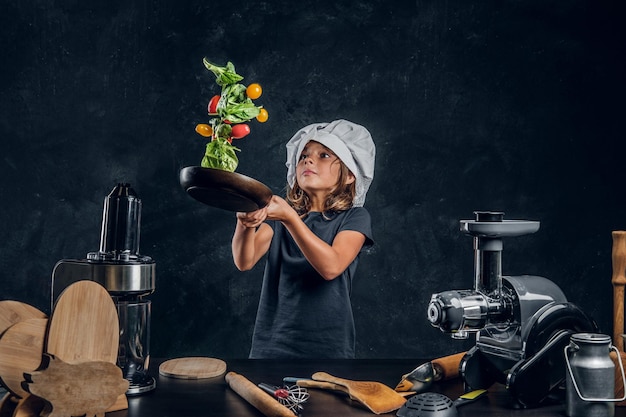 Całkiem mała dziewczynka rzuca warzywa na patelnię w ciemnym studiu fotograficznym.