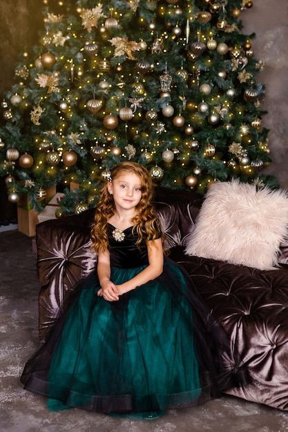 Całkiem mała dziewczynka siedzi obok choinki. Mała księżniczka w fantazyjnej sukience, długie kręcone włosy, motyw noworoczny.