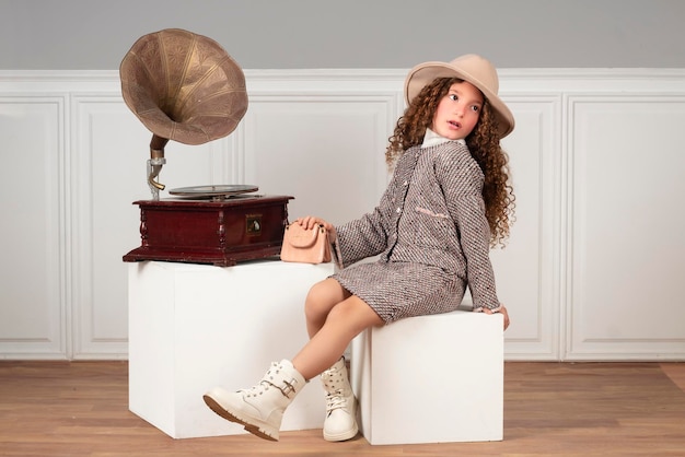 Całkiem mała dziewczynka w klasycznym stroju siedzi obok gramofonu.