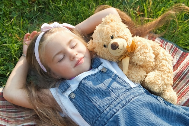 Całkiem małe dziecko dziewczynka z zamkniętymi oczami ustanawiające z jej zabawką pluszowego misia na kocu na zielonej trawie w lecie drzemał.